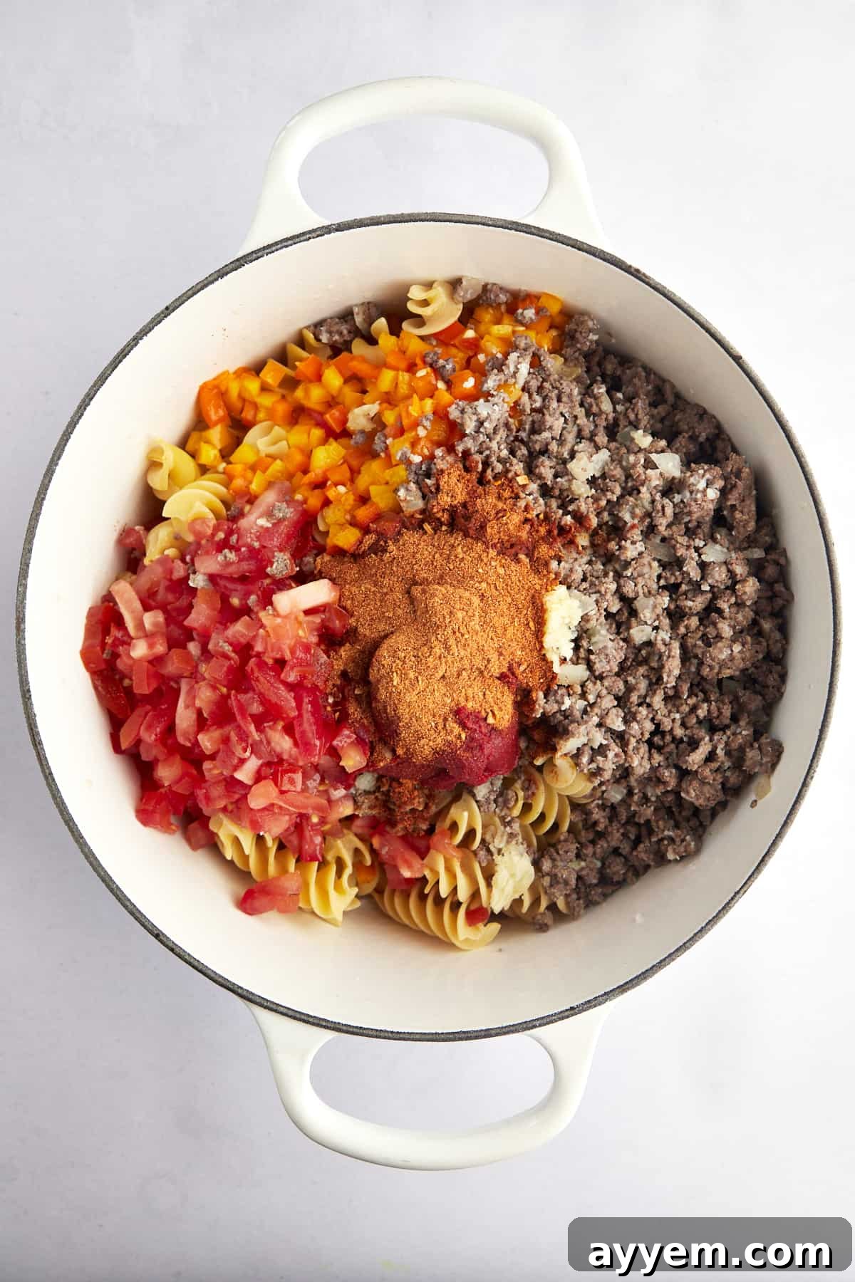 Overhead image of cooked beef, bell peppers, tomatoes, raw noodles, and taco seasonings in a pot. 