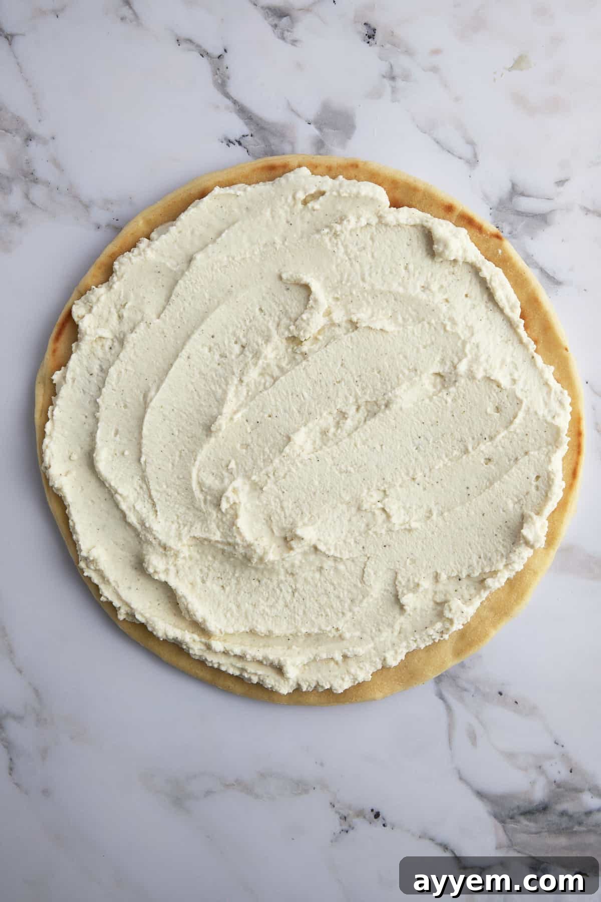 Pizza crust generously spread with a smooth, creamy layer of whipped feta, forming the unique white sauce base for the gyro pizza.