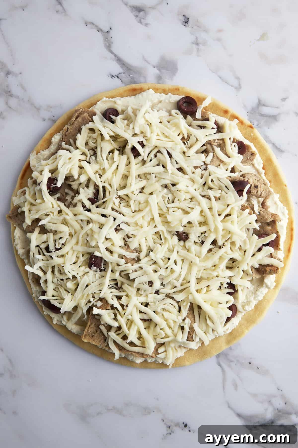 Fully assembled pizza ready for baking, topped with whipped feta, gyro meat, olives, and a final layer of shredded Mozzarella cheese.