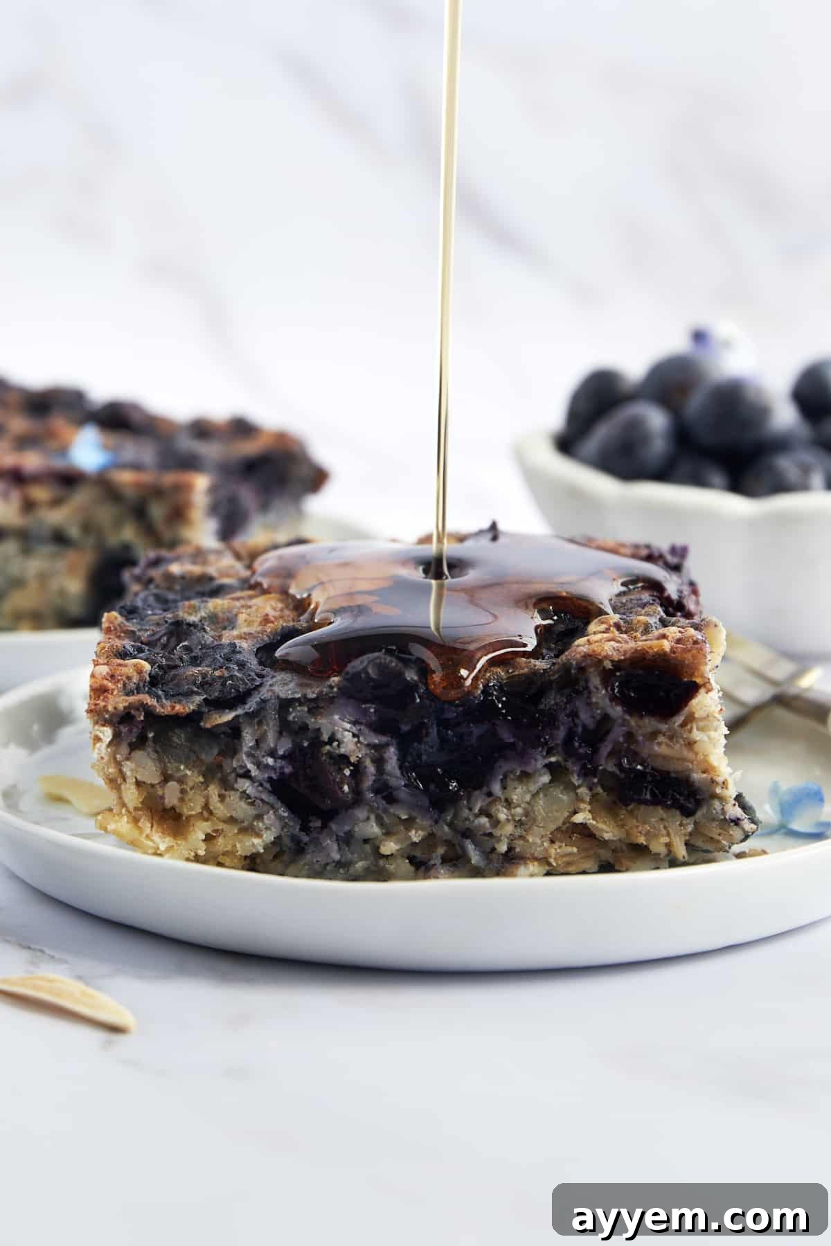 A generous serving of warm blueberry baked oatmeal, beautifully garnished with fresh blueberries and a golden stream of maple syrup being poured over the top, ready to be enjoyed.