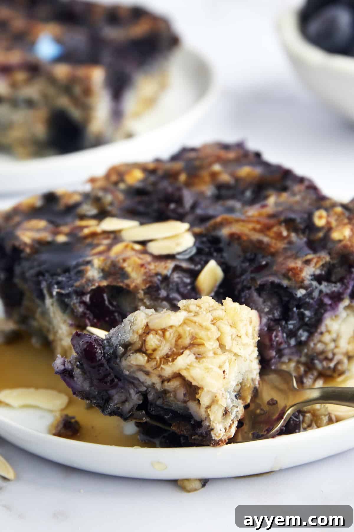 A close-up shot of a plate with a generous slice of blueberry baked oats, showing its tender interior and juicy blueberries. A fork is poised, holding a perfect bite, inviting you to taste.