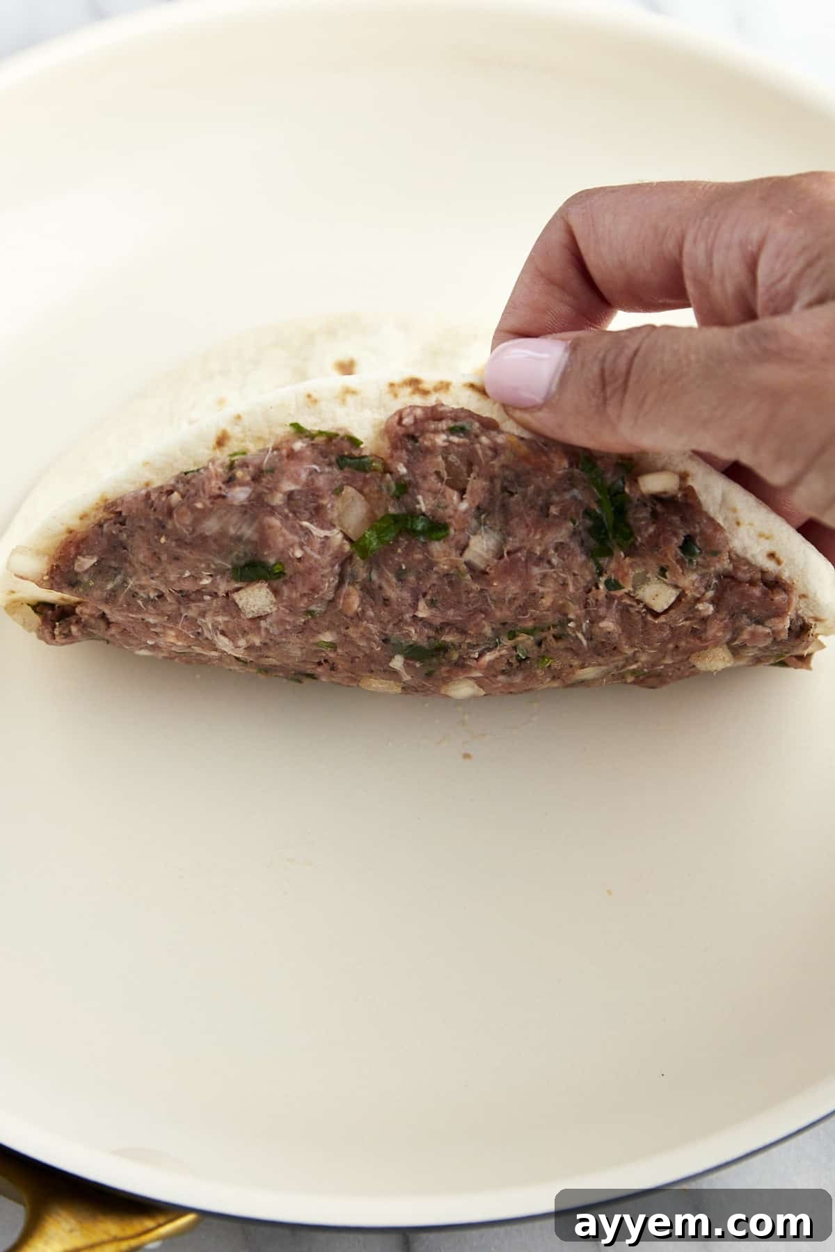 A hand placing a tortilla with seasoned ground beef on it into a pan. 