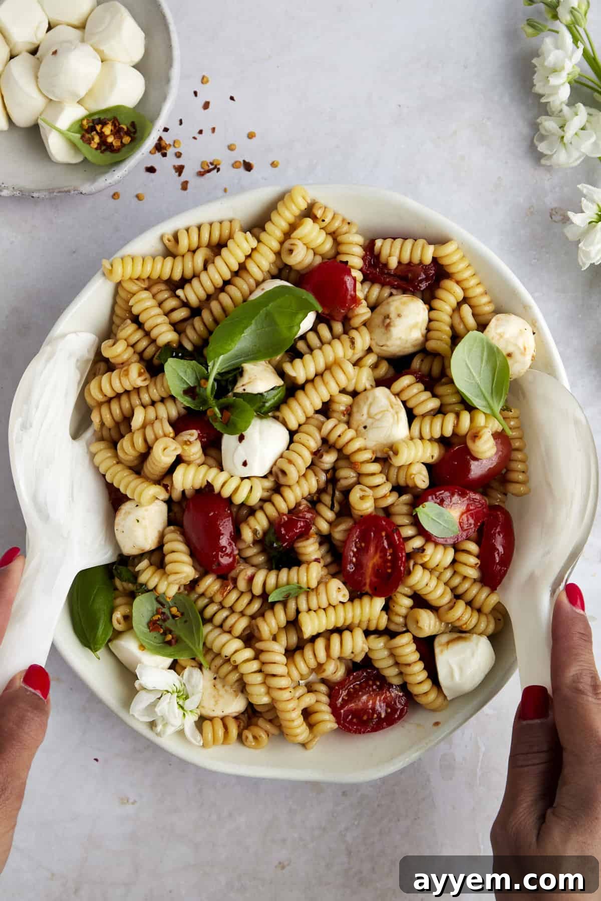Two hands gently tossing Caprese pasta salad in a large glass bowl, showing the mix of pasta, tomatoes, mozzarella, and basil.