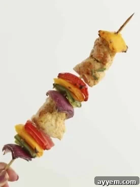 a hand holding an oven-baked chipotle chicken skewer with red, yellow, and green peppers and red onion