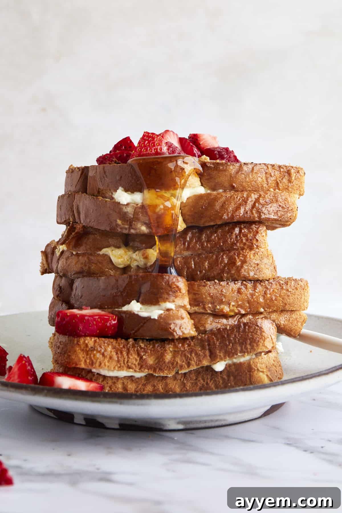 Air fryer french toast stuffed with cream cheese stacked on a plate topped with strawberries and syrup. 