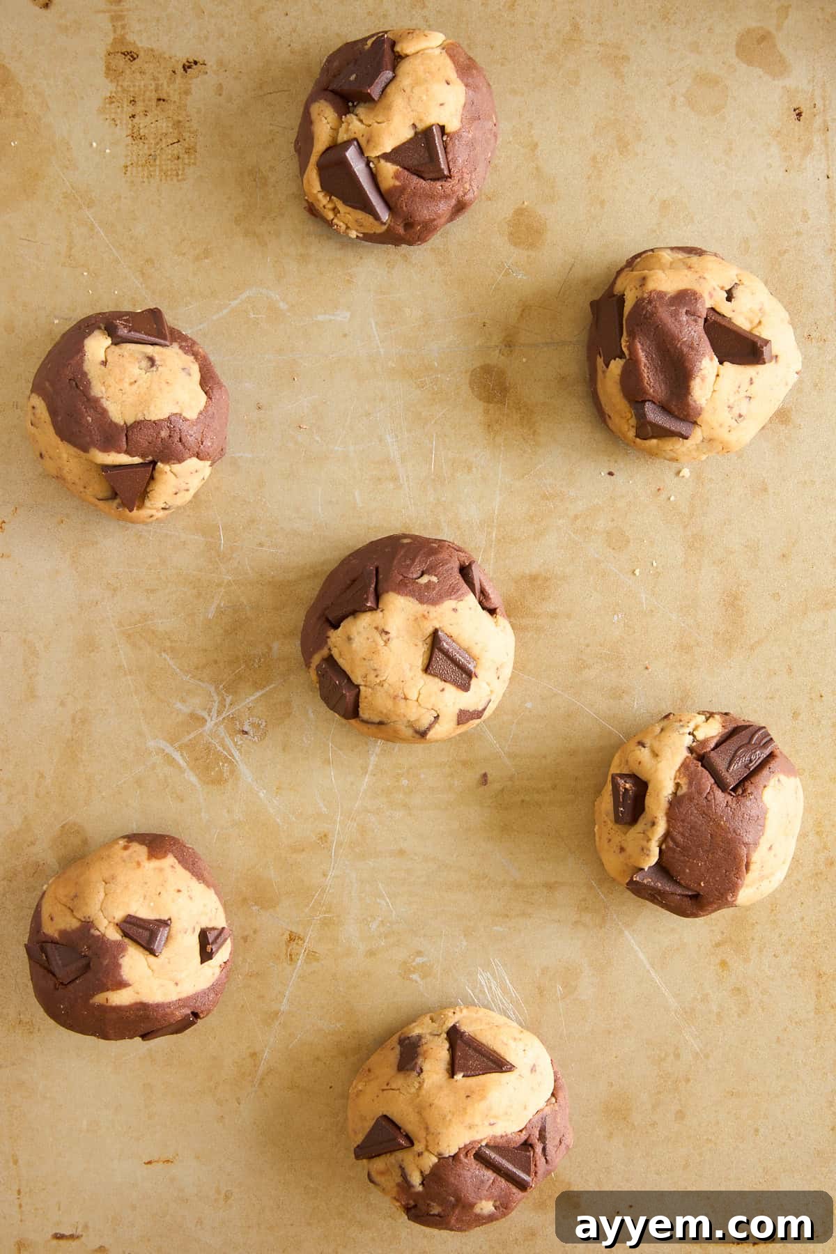 Seven unbaked, swirled chocolate and vanilla cookie dough balls with chocolate chunks are arranged on a lightly scratched baking sheet.