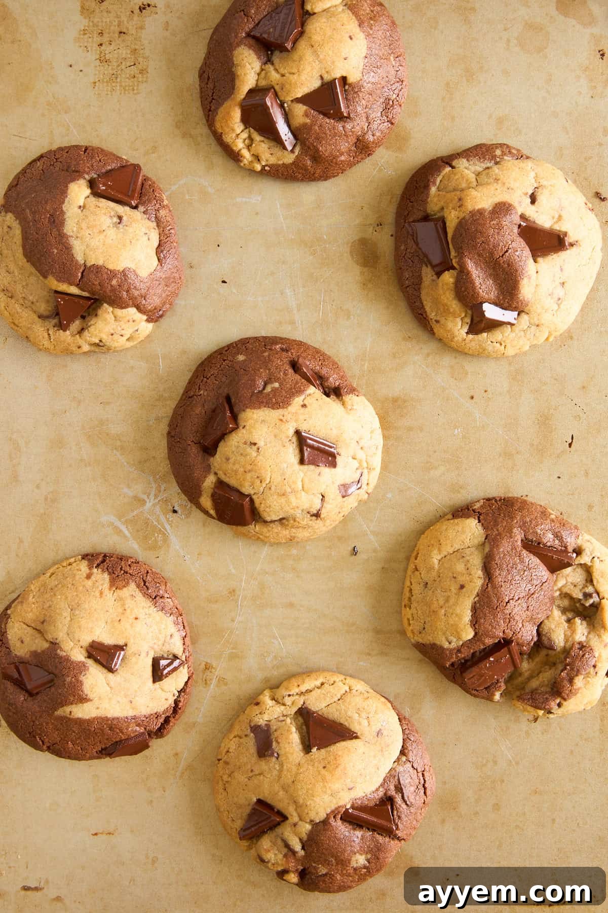 Seven large, marble-patterned cookies with chunks of chocolate are arranged on a tan baking sheet. The cookies have swirls of light and dark dough, and the surface is slightly cracked.