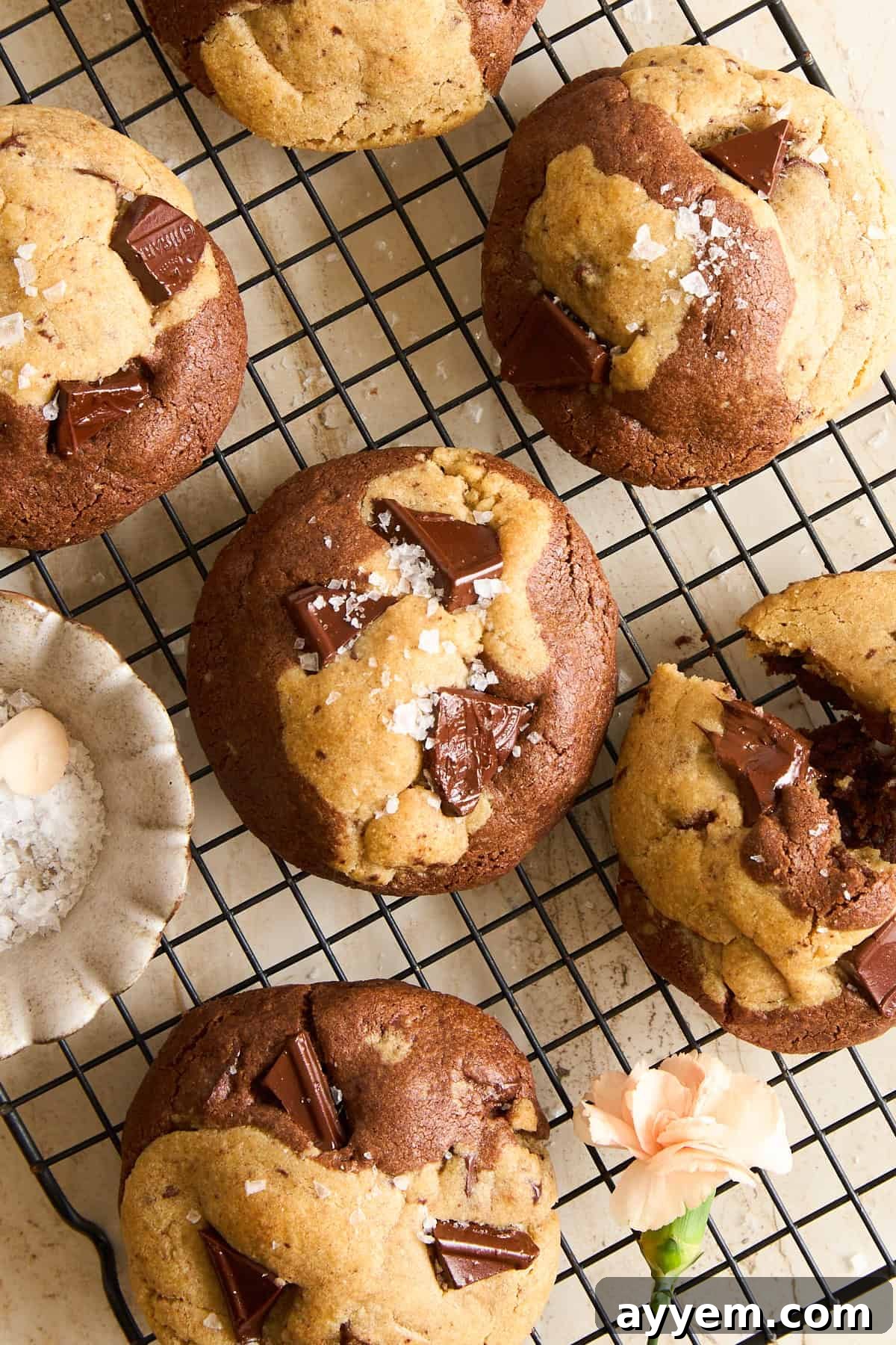 Chocolate chip cookies with sea salt flakes rest on a cooling rack. Some cookies have visible chocolate chunks, and a small bowl of sea salt and a light peach flower sit nearby on the beige surface.