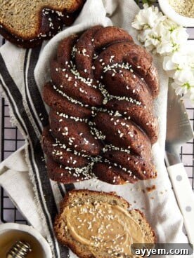A freshly baked loaf of tahini banana bread, drizzled with honey and sprinkled with black and white sesame seeds, with a slice removed, topped with nut butter.