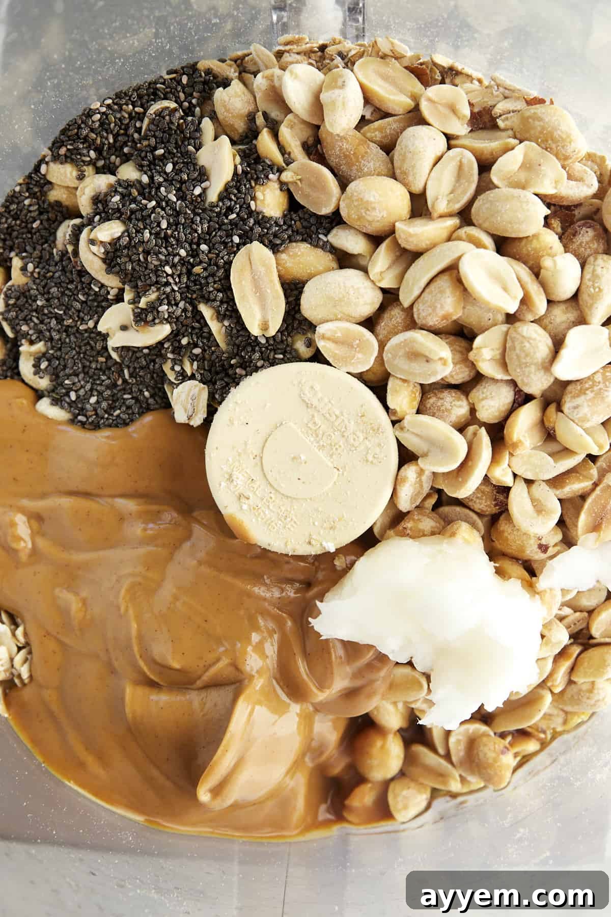 Raw peanuts, chia seeds, a spoonful of peanut butter, and solid coconut oil placed around a food processor bowl before blending.