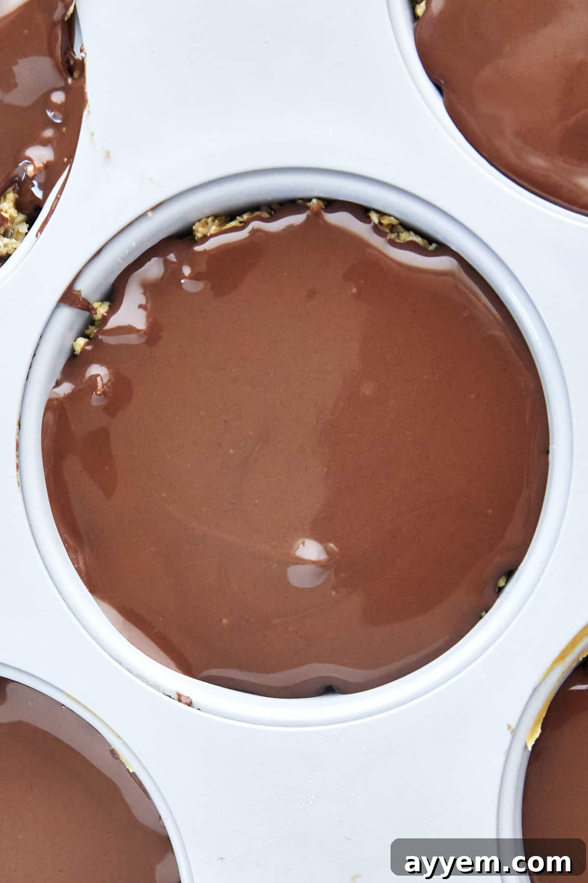 Melted chocolate being poured over a no-bake peanut butter oat cup in a muffin pan, creating the top layer.
