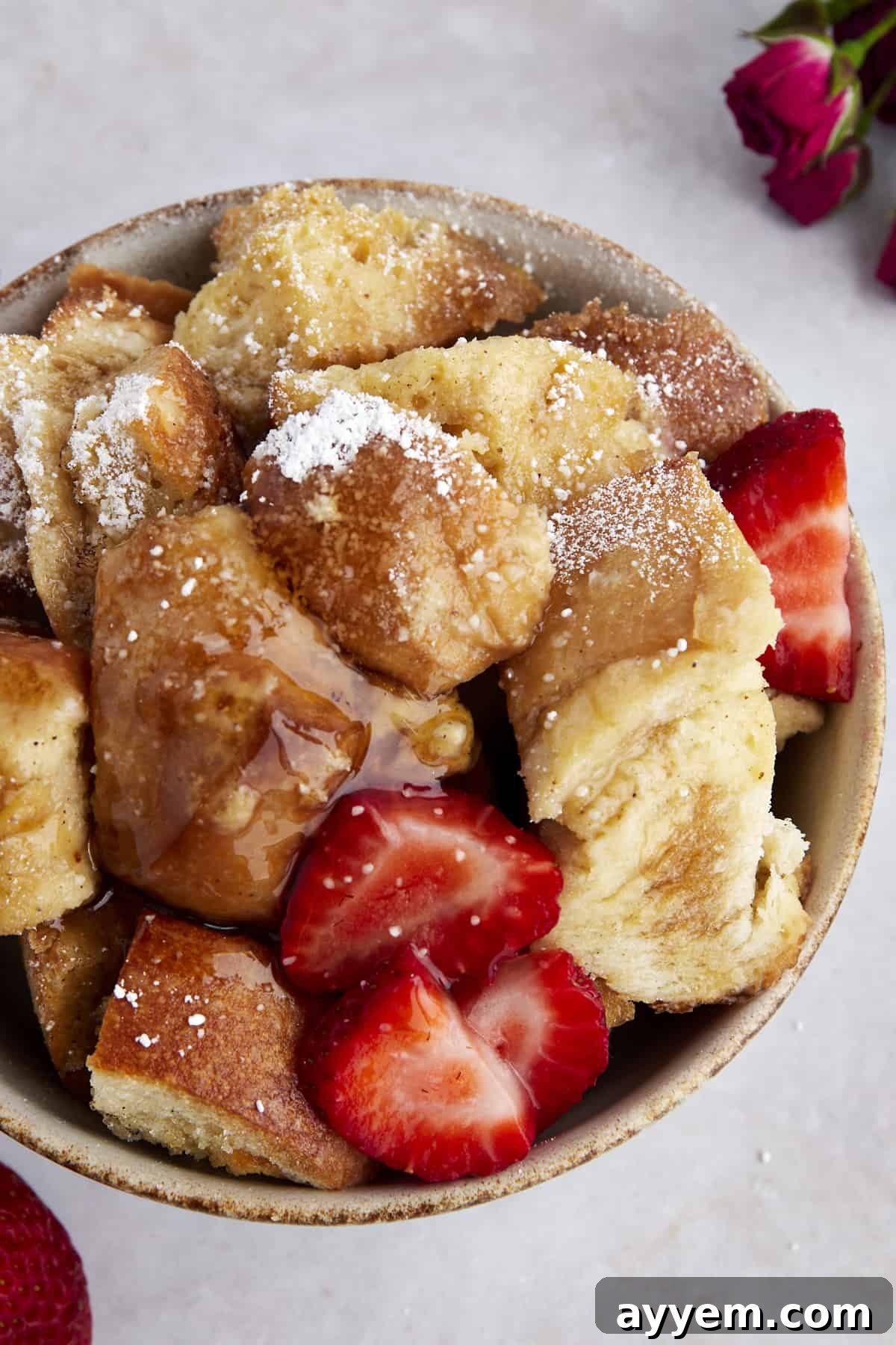 A bowl of crockpot French toast casserole topped with sliced strawberries, powdered sugar, and maple syrup. 