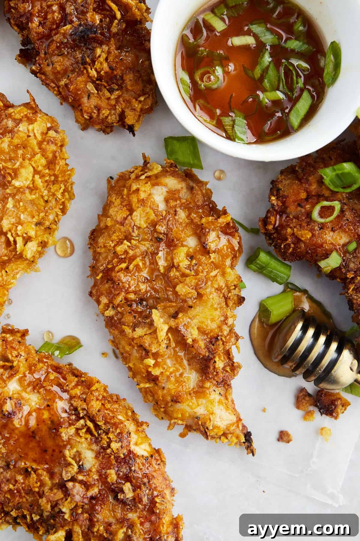 Golden brown, crispy baked cornflake chicken tenders generously drizzled with glistening hot honey, resting on a white platter. A small bowl of extra hot honey is visible in the background, inviting diners to dip.