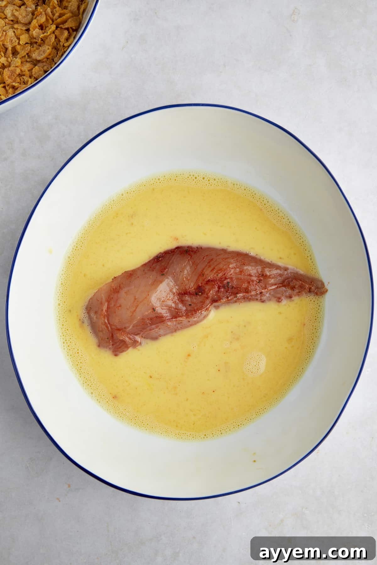 Chicken tenders seasoned and submerged in an egg wash, preparing for coating. The chicken looks moist and ready to absorb the next layer of flavor.