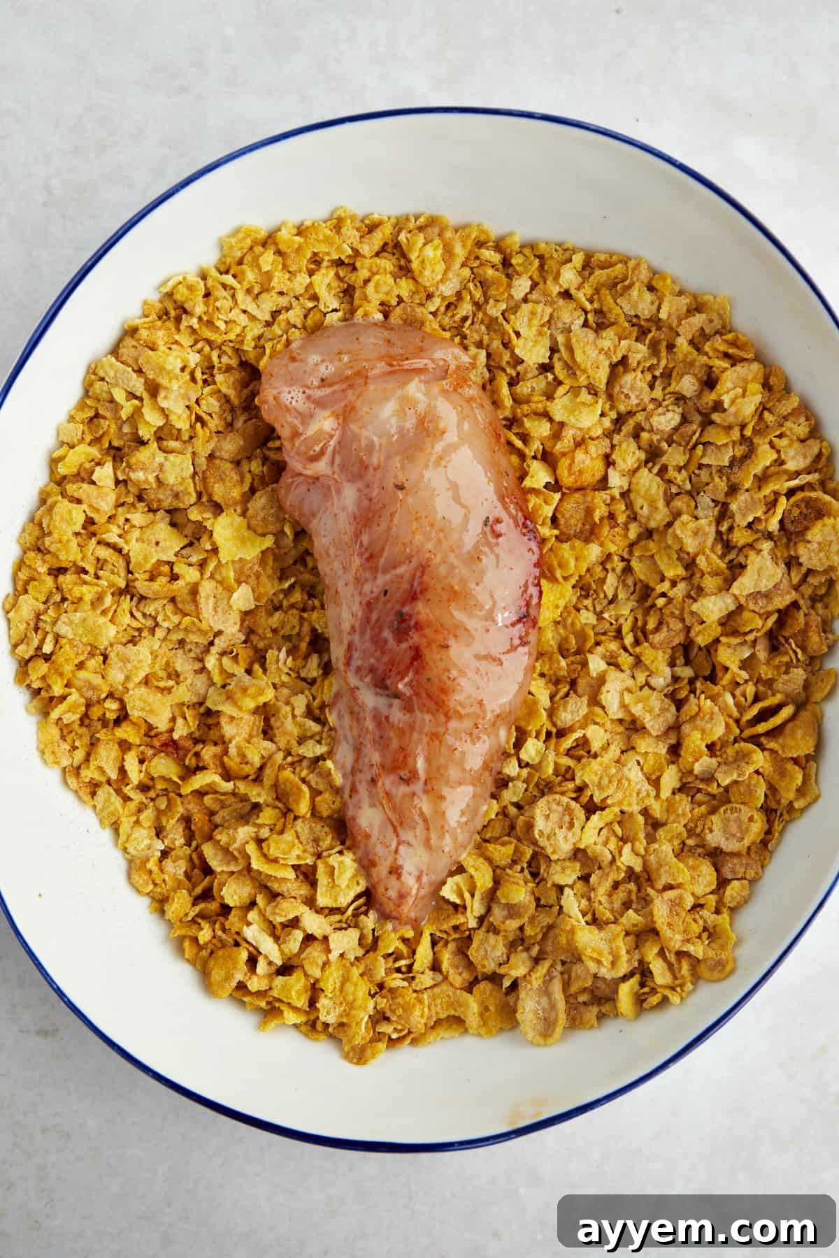 Raw chicken tender, perfectly coated in a coarse mixture of crushed cornflakes and spices, resting in a bowl, showcasing the ideal breading texture.