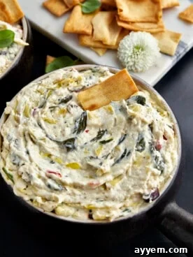 A bubbling, warm bowl of Mediterranean-inspired crockpot spinach and artichoke dip, with a pita chip ready for dipping.