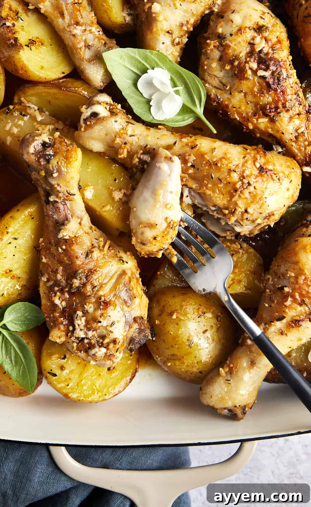 A fork holding a juicy bite of chicken and potato, with a full dish of garlic parmesan drumsticks and roasted potatoes in the background.