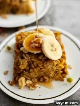 A piece of baked pumpkin oatmeal on a white plate topped with walnuts and banana coins with a drizzle of maple syrup being poured on top.