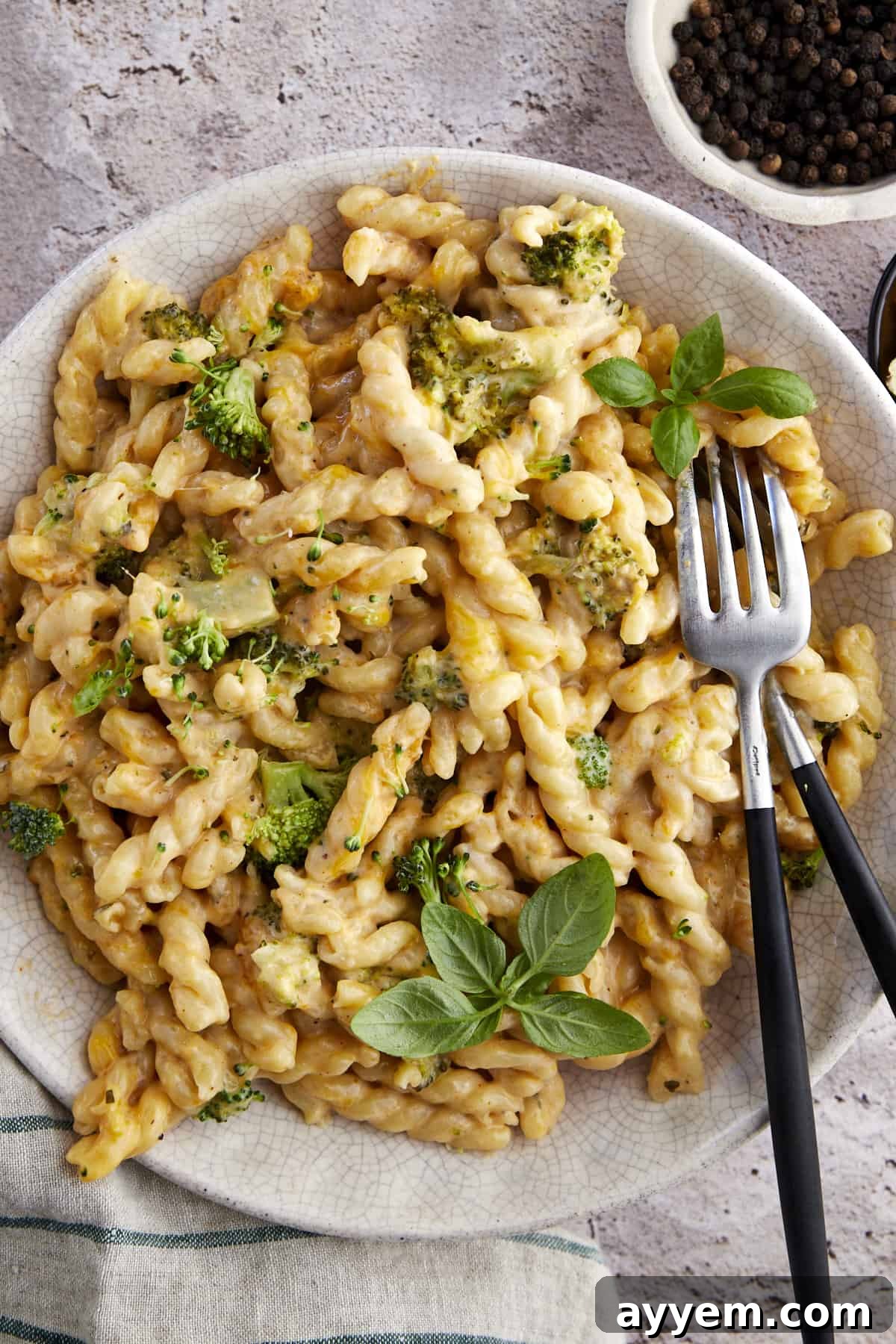 A golden-brown baked broccoli mac and cheese in a rustic ceramic bowl, with two forks invitingly placed, ready to be served. The cheese is bubbly and slightly browned on top, with visible broccoli florets and pasta.