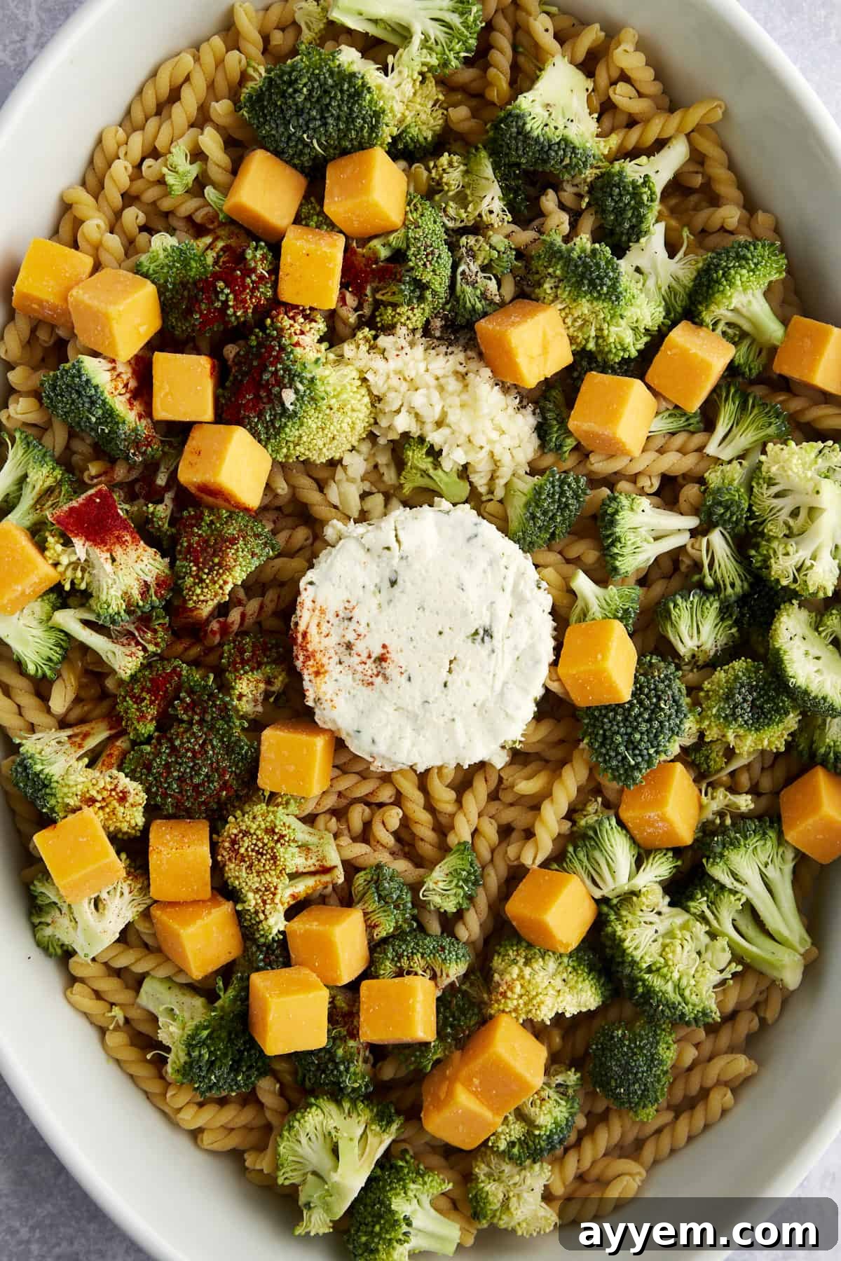 Overhead shot showcasing a large baking dish with uncooked Gemelli pasta, a block of Boursin cheese, cubed cheddar, fresh garlic, and broccoli florets, all prepped before adding liquid.