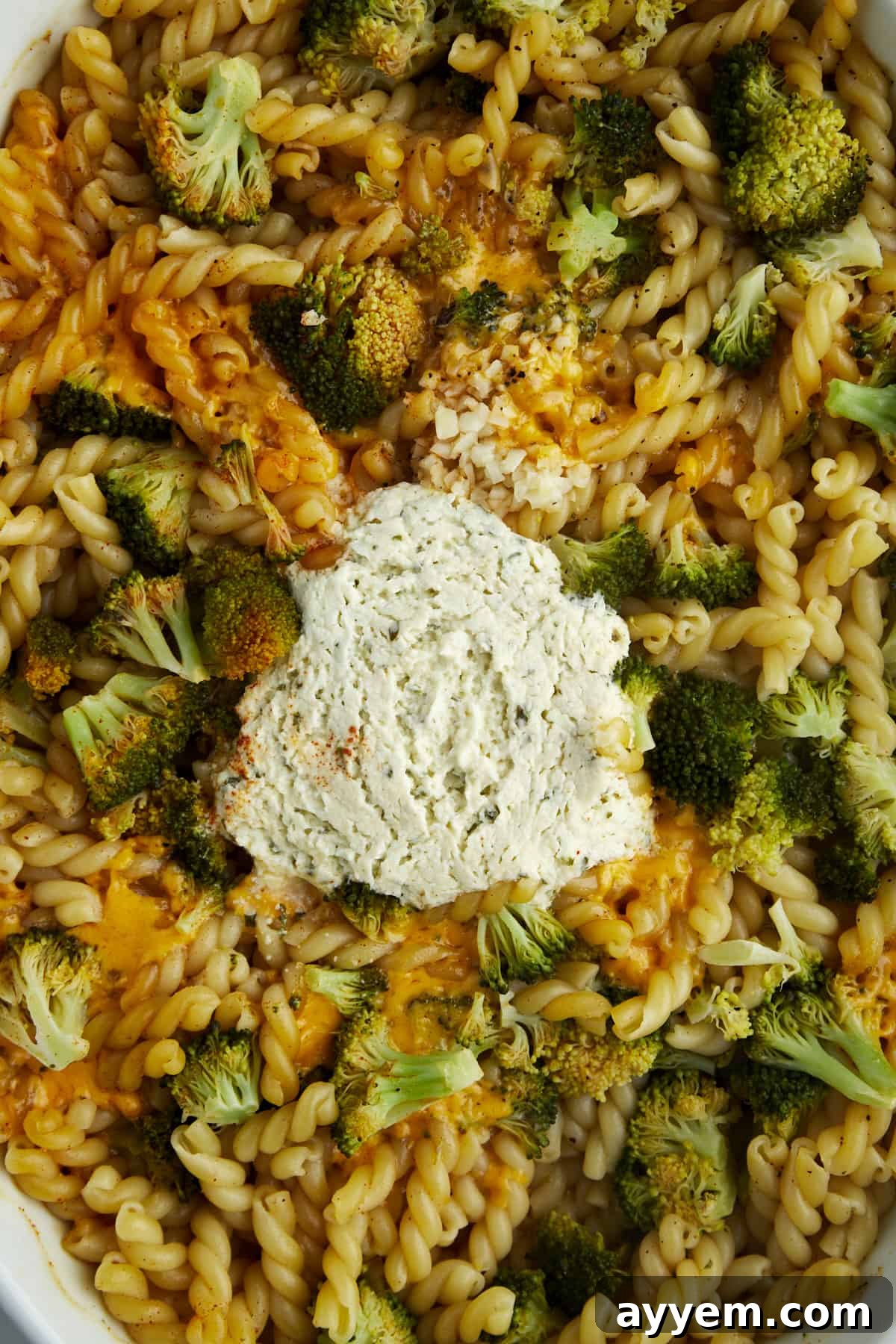 A close-up overhead view of the ingredients in a baking dish, featuring uncooked pasta, a block of Boursin cheese, chopped broccoli, and cubed cheddar, waiting for the liquid base.