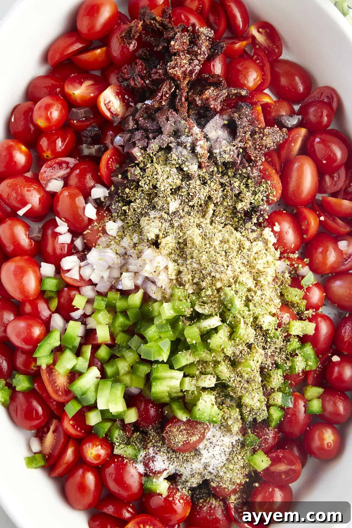 A baking dish filled with vibrant, uncooked Mediterranean ingredients: tomatoes, olives, bell pepper, onion, garlic, and seasonings, ready for the oven.