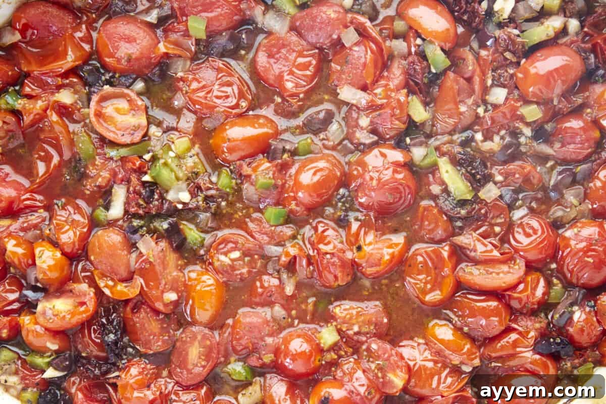 A close-up view of roasted baby tomatoes, Kalamata olives, and sliced bell peppers in a baking dish, glistening with olive oil and herbs after being baked.