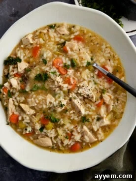 A steaming bowl of creamy chicken and rice soup, with chunks of chicken and vegetables, and a spoon for serving.