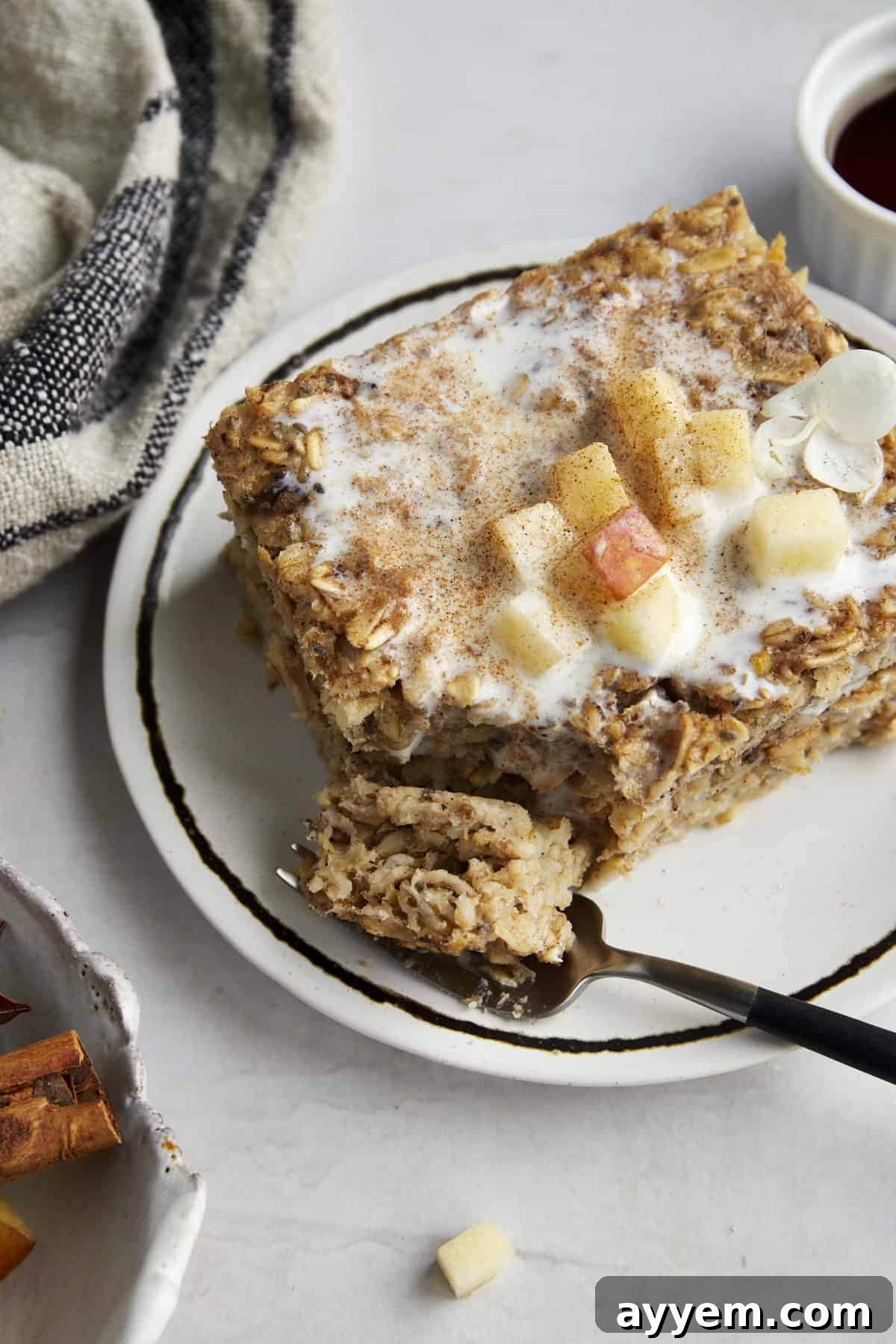 A slice of baked apple oatmeal on a plate topped with apples and milk with a fork holding a bite. 
