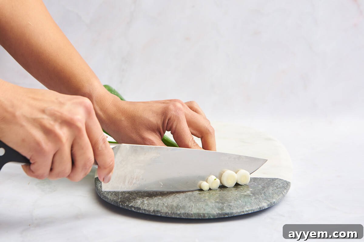 Two hands meticulously chopping the white ends of green onions into small, uniform pieces on a clean cutting board.