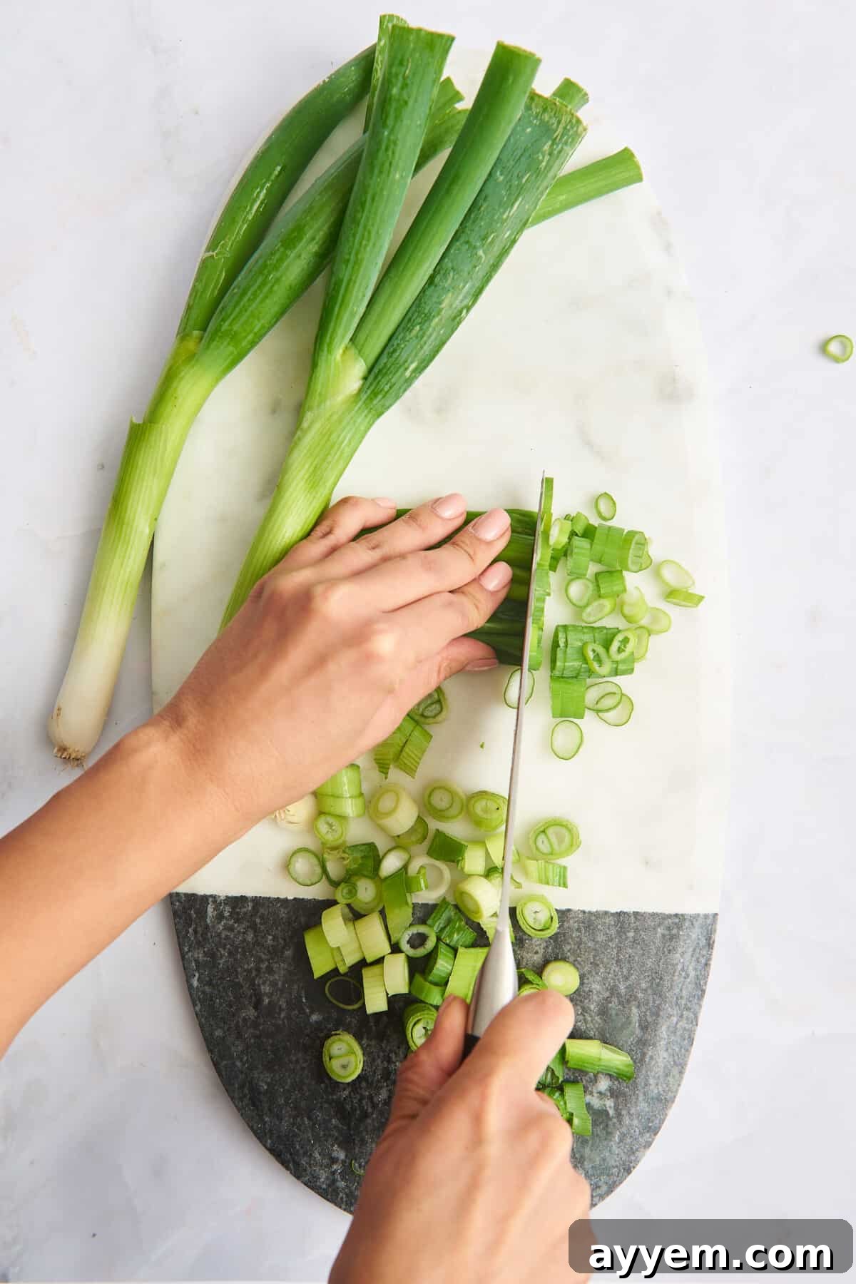 A cutting board showcasing whole and finely chopped green onions, with a hand actively slicing scallions, demonstrating the cutting process.