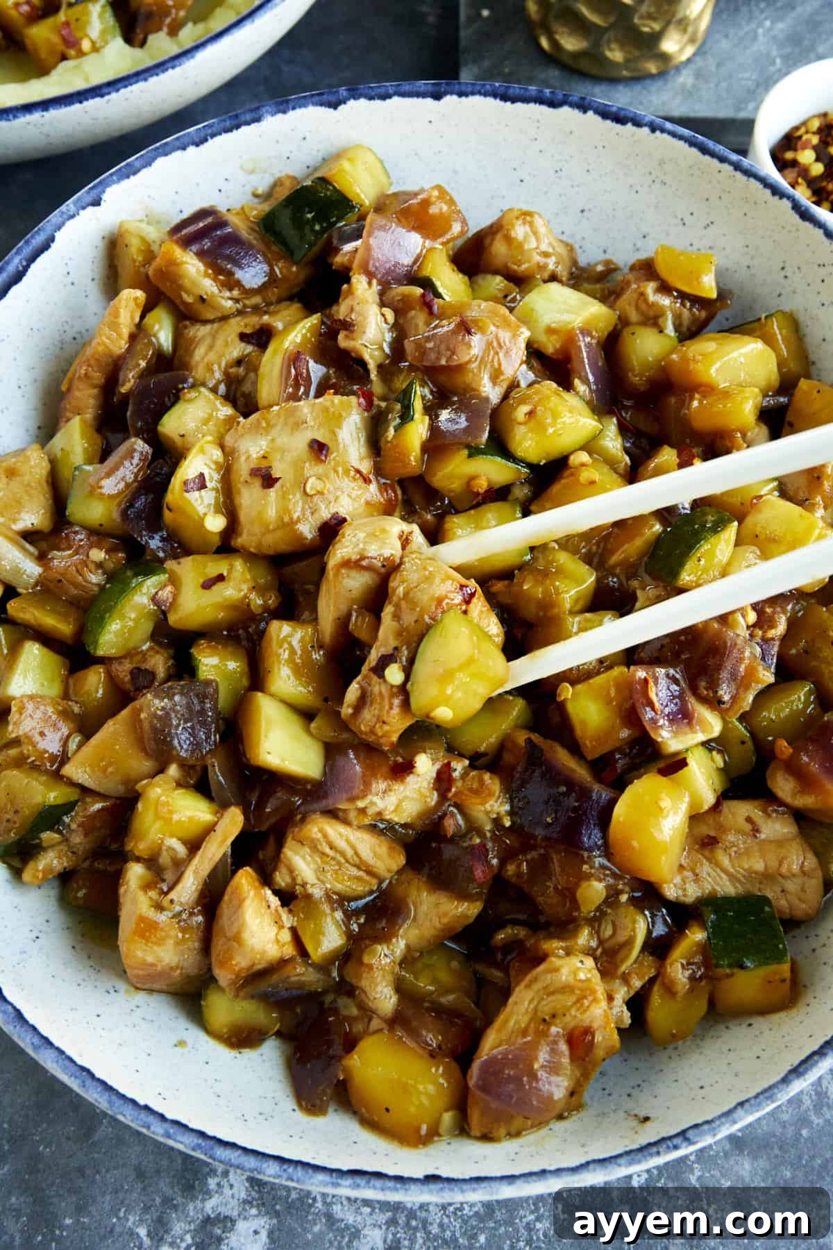 Two chopsticks gracefully picking up a succulent piece of chicken and tender zucchini from a stir-fry dish.