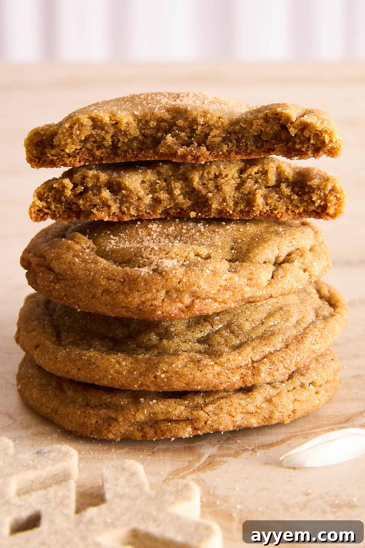 A stack of five brown sugar cookies, with the top cookie broken in half to show a soft, chewy texture inside. The cookies are sprinkled with sugar and rest on a light wooden surface.