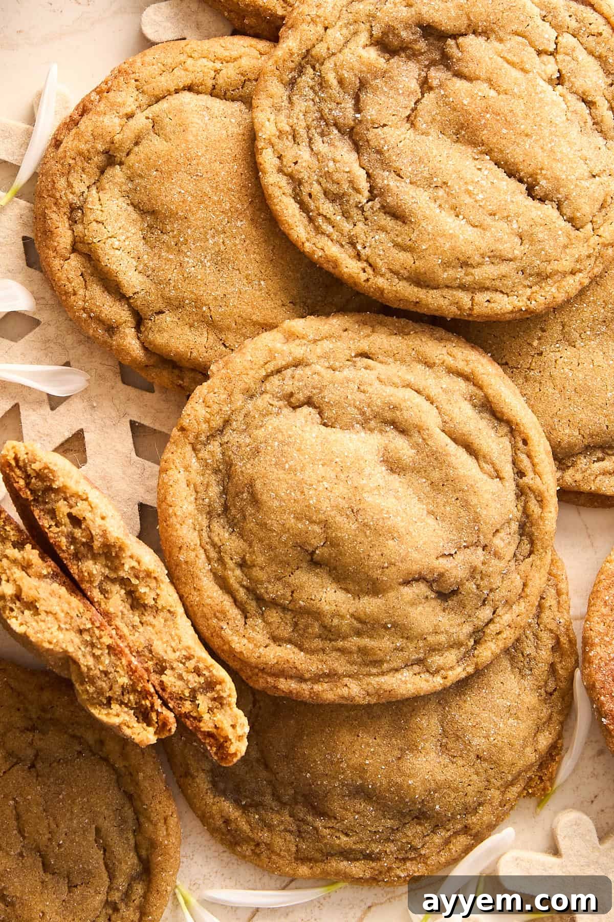 A close-up of several golden brown cookies, some stacked and one cookie broken in half, revealing a soft, chewy interior. The cookies have a slightly crackled surface and are placed on a light-colored surface.