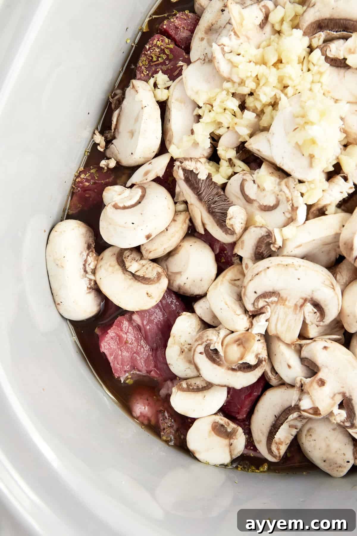 Uncooked beef, sliced mushrooms, minced garlic, and sauce ingredients nestled in a slow cooker, before cooking.