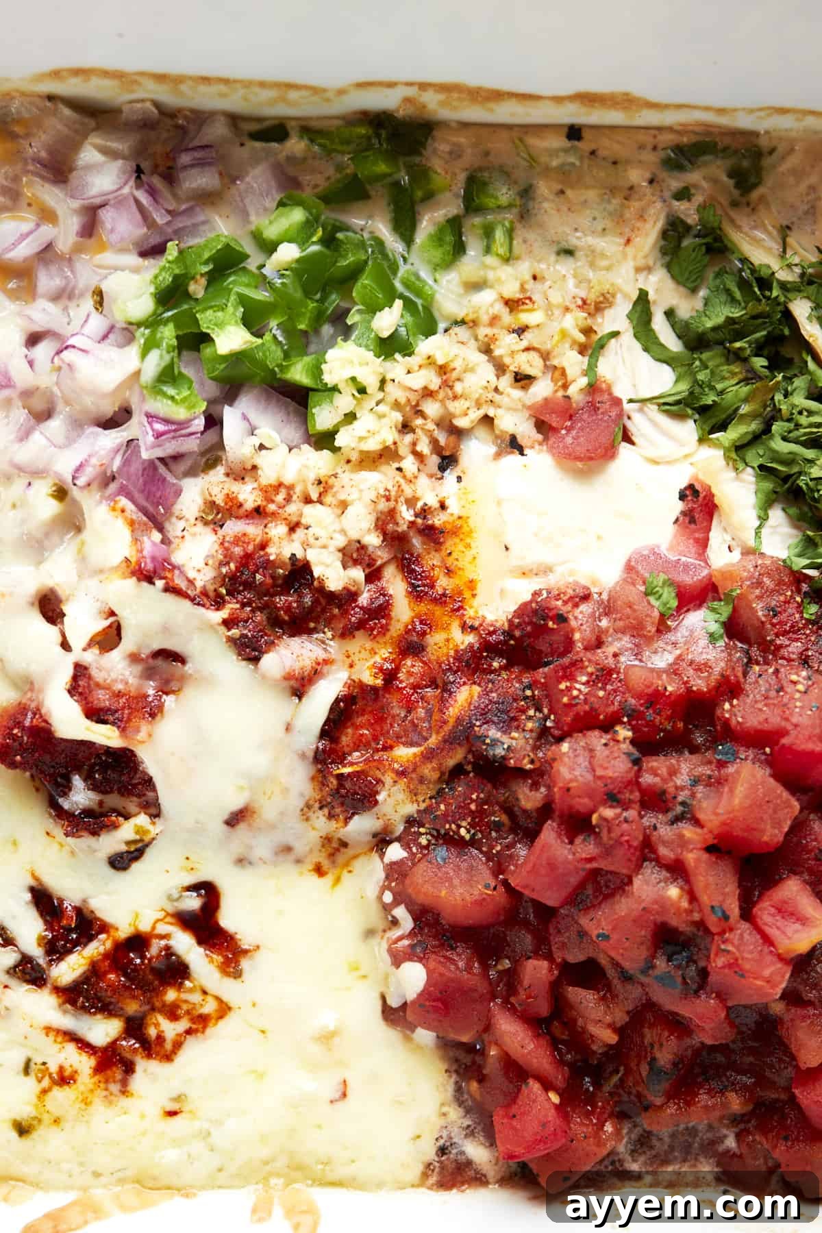 Baked chicken queso ingredients in a baking dish topped with evaporated milk, showcasing the creamy liquid settling among the cheeses and other components before entering the oven.