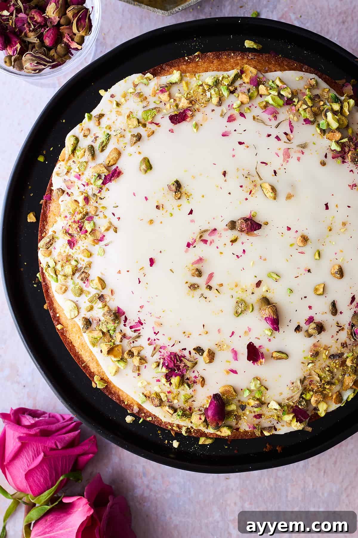 A beautifully decorated Persian love cake, generously topped with glistening orange glaze, delicate pink rose petals, and vibrant green crushed pistachios, ready to be served.