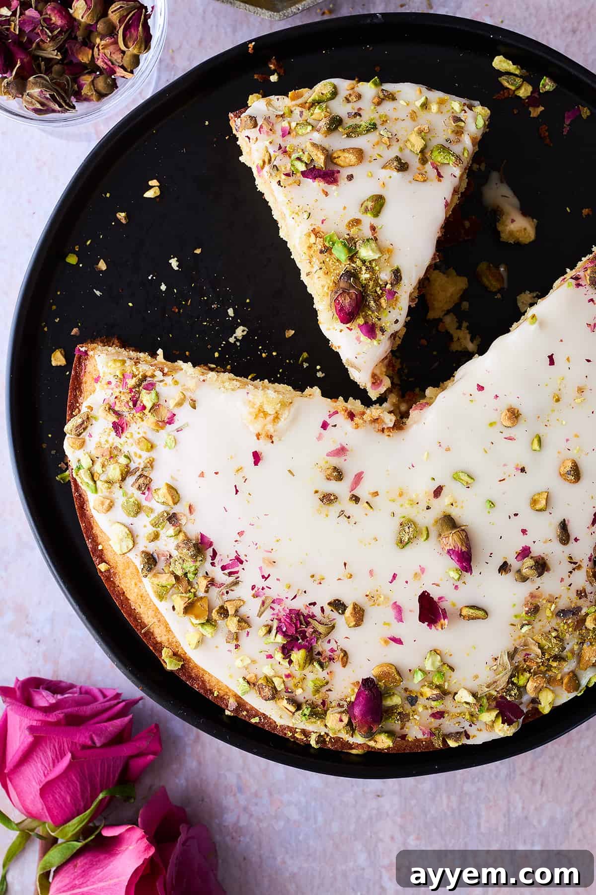 A large Persian love cake, with several pieces already cut out, and one perfect slice resting on its side, showcasing its moist texture and vibrant toppings.