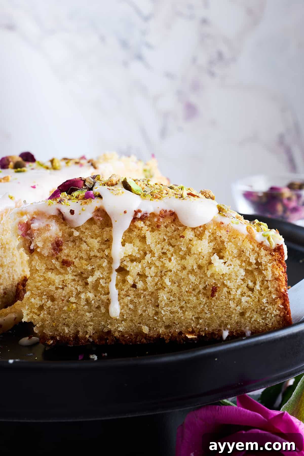 A close-up side view of a perfectly sliced Persian love cake, showcasing its moist, fluffy interior, the luscious orange glaze, and the vibrant toppings of pistachios and rose petals.