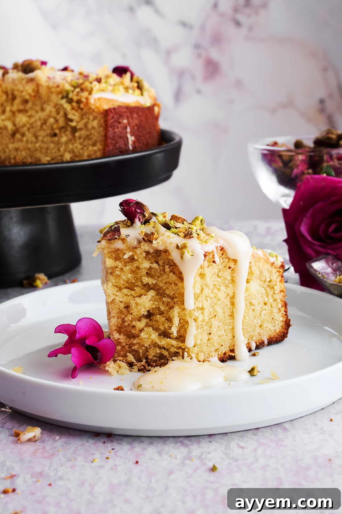 A close-up of the Persian love cake's textured surface, with bright green pistachios and delicate pink rose petals scattered over the creamy orange glaze.