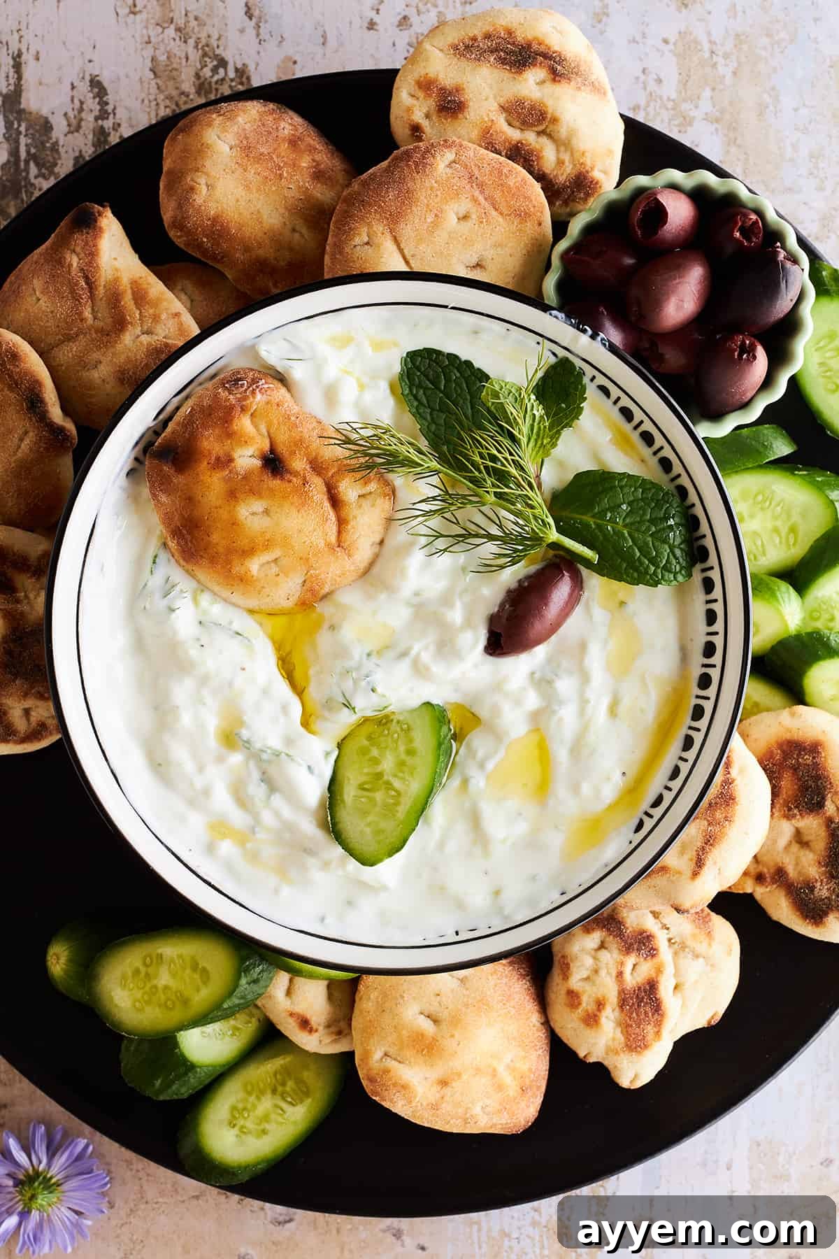 A bowl of tzatziki sauce on a plate with pita, olives, and cucumber slices. 