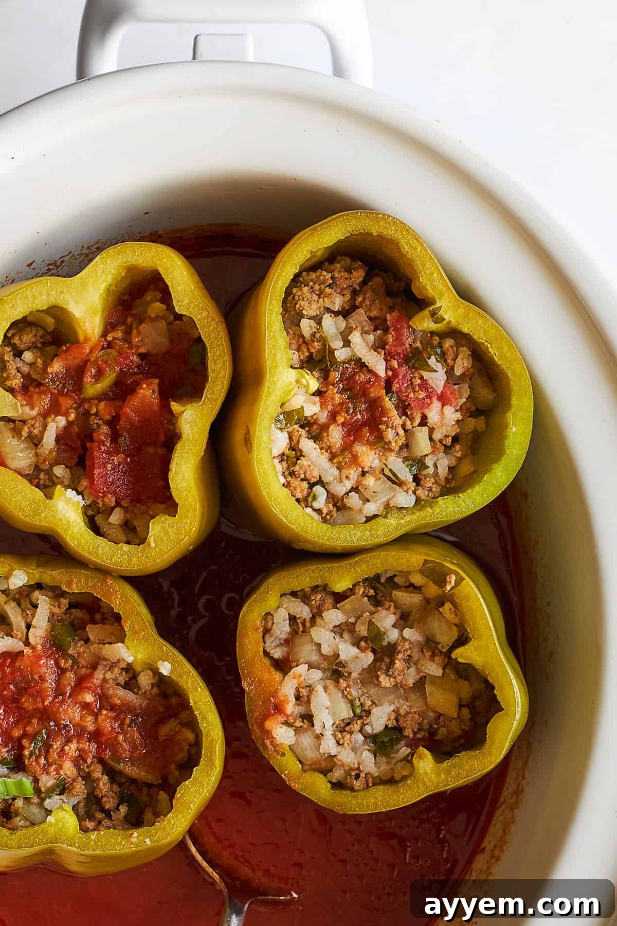 Stuffed peppers with rice simmering in a slow cooker, with a spoon stirring the tomato sauce base.