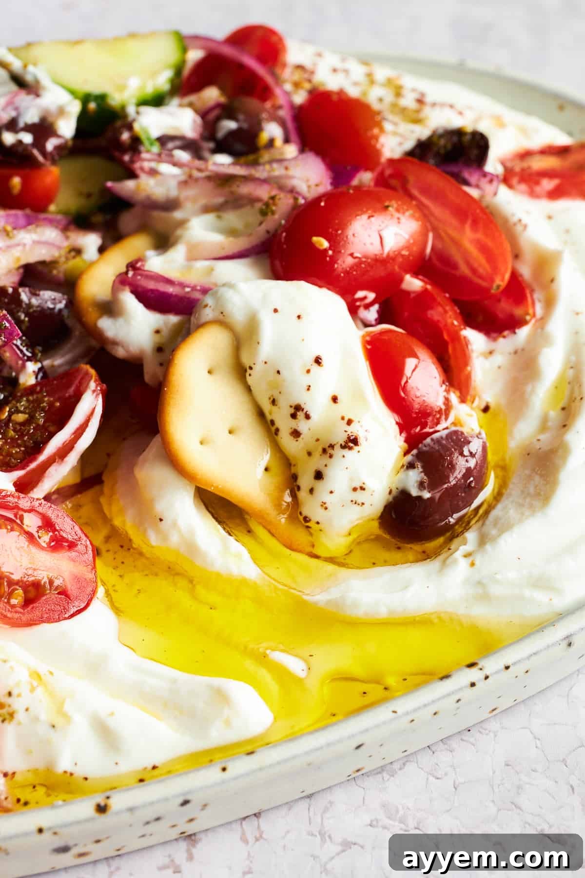 A crisp, golden cracker being dipped into a bowl of creamy Greek salad dip, highlighting the vibrant layers of fresh vegetables.
