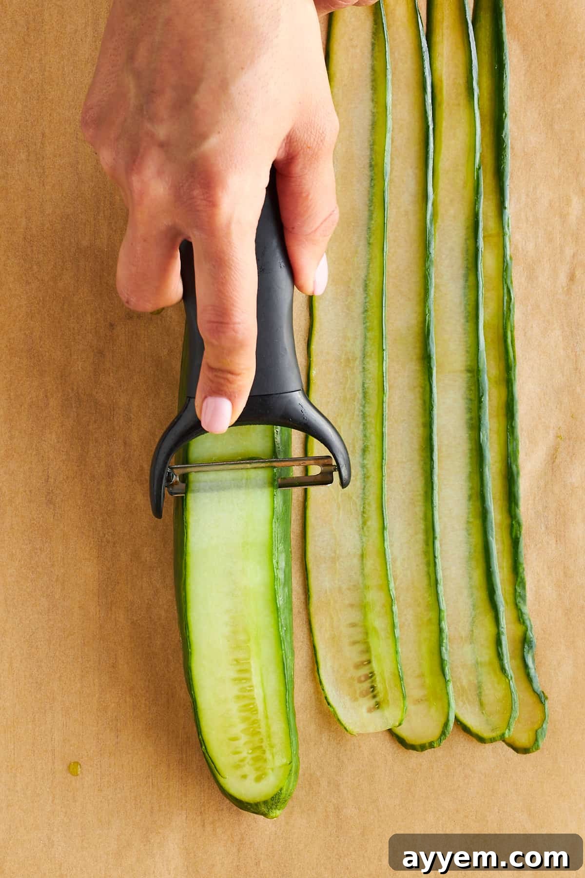 A hand skillfully slicing an English cucumber into thin, elegant strips using a vegetable peeler.