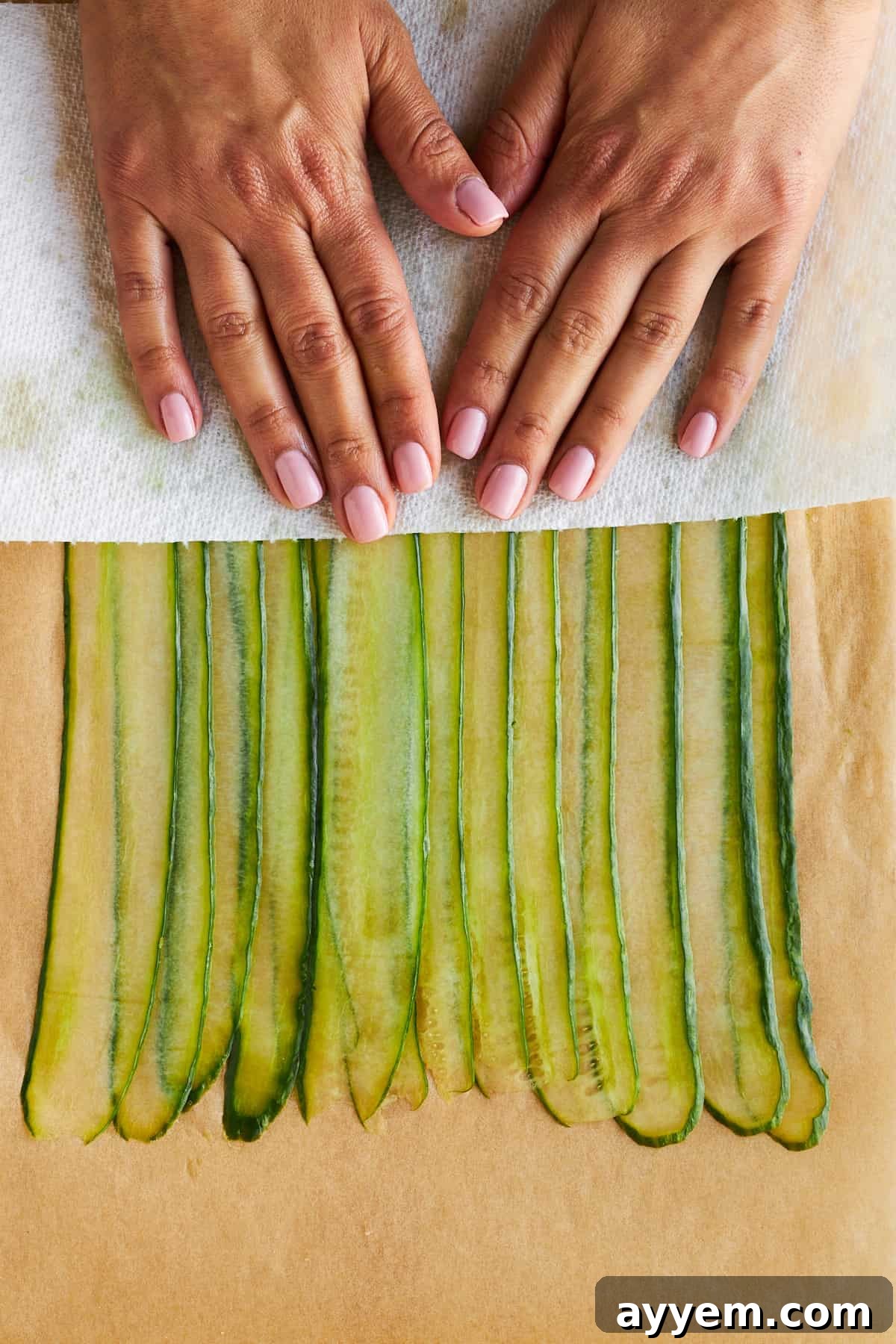Hands gently patting layered cucumber strips dry with a paper towel, crucial for preventing sogginess and ensuring tight rolls.