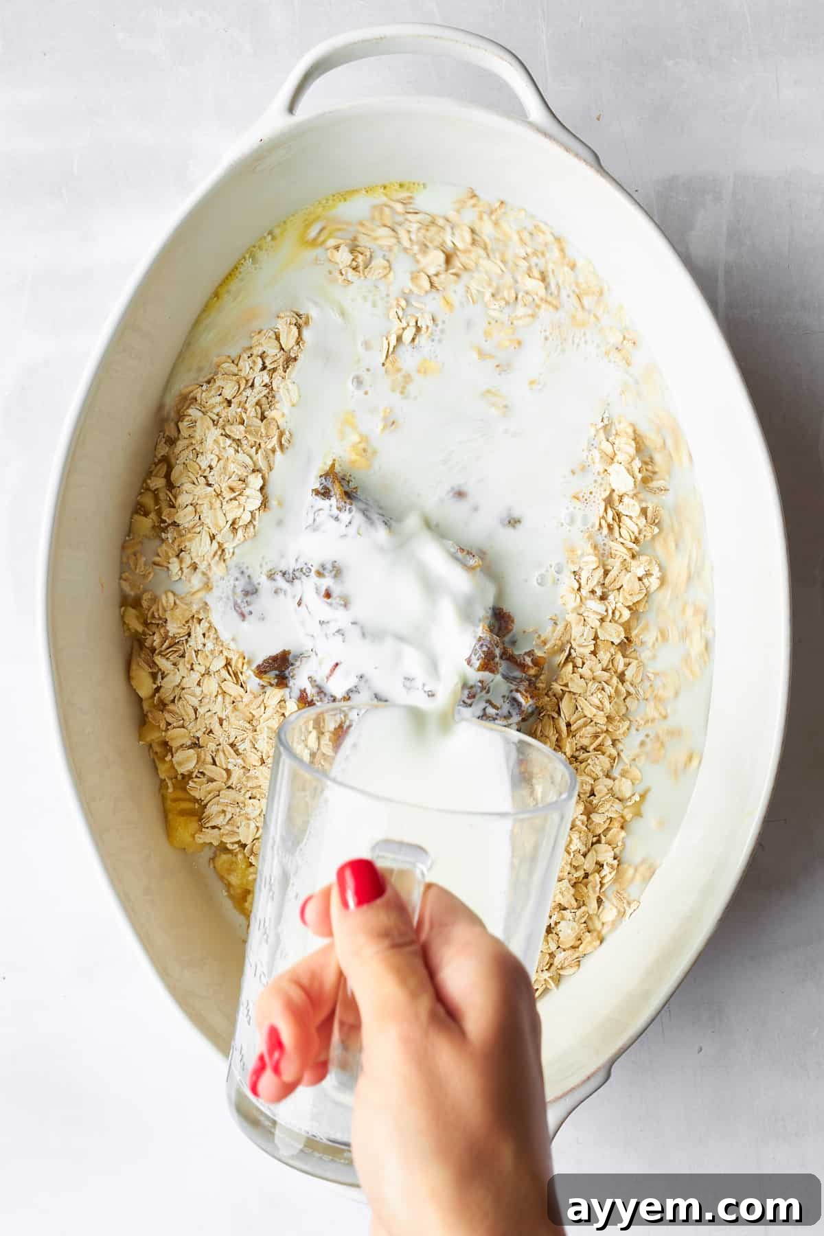 Milk being poured over the banana date oatmeal bake ingredients in an oval baking dish.