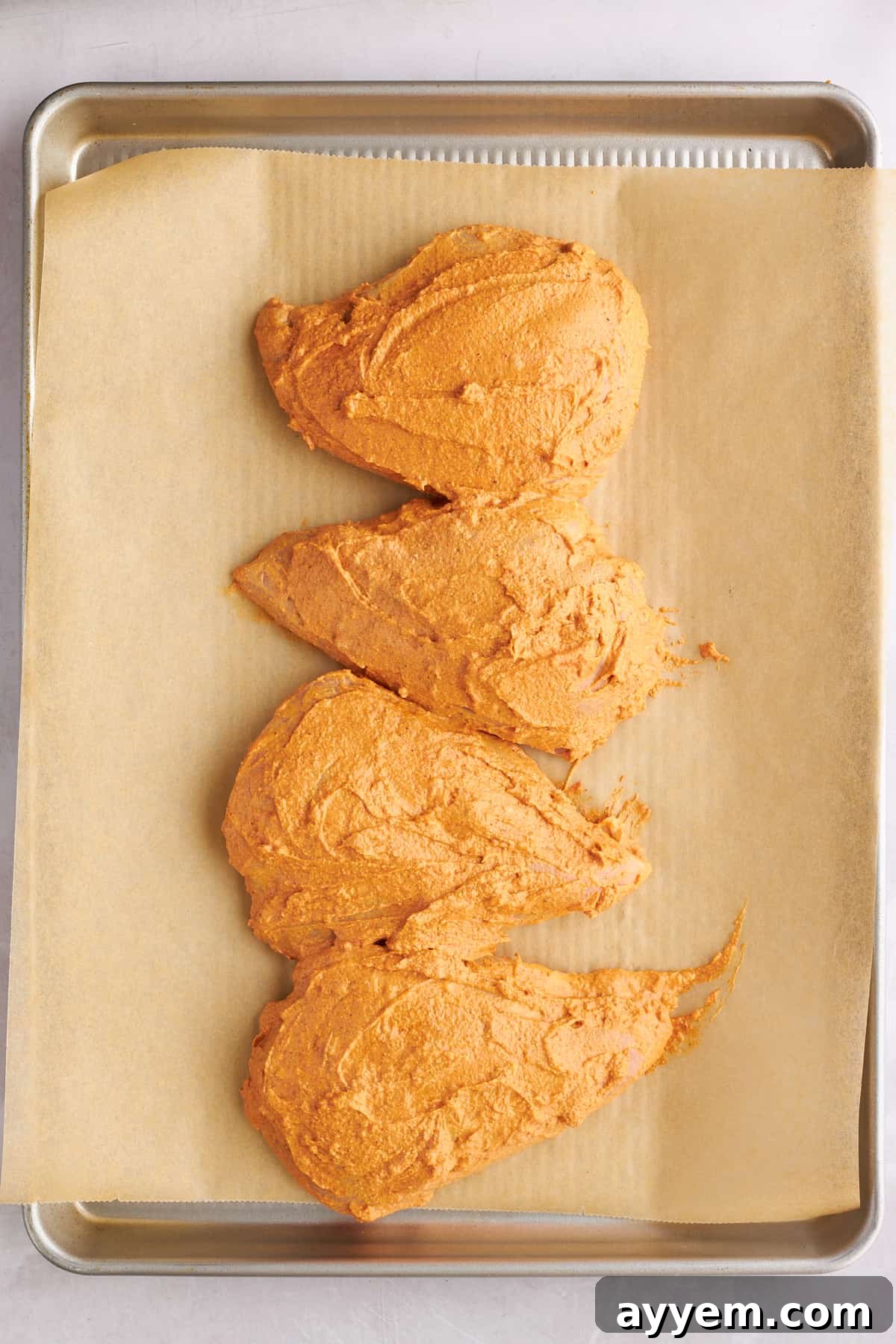 Four marinaded chicken breasts on a parchment paper-lined baking sheet.