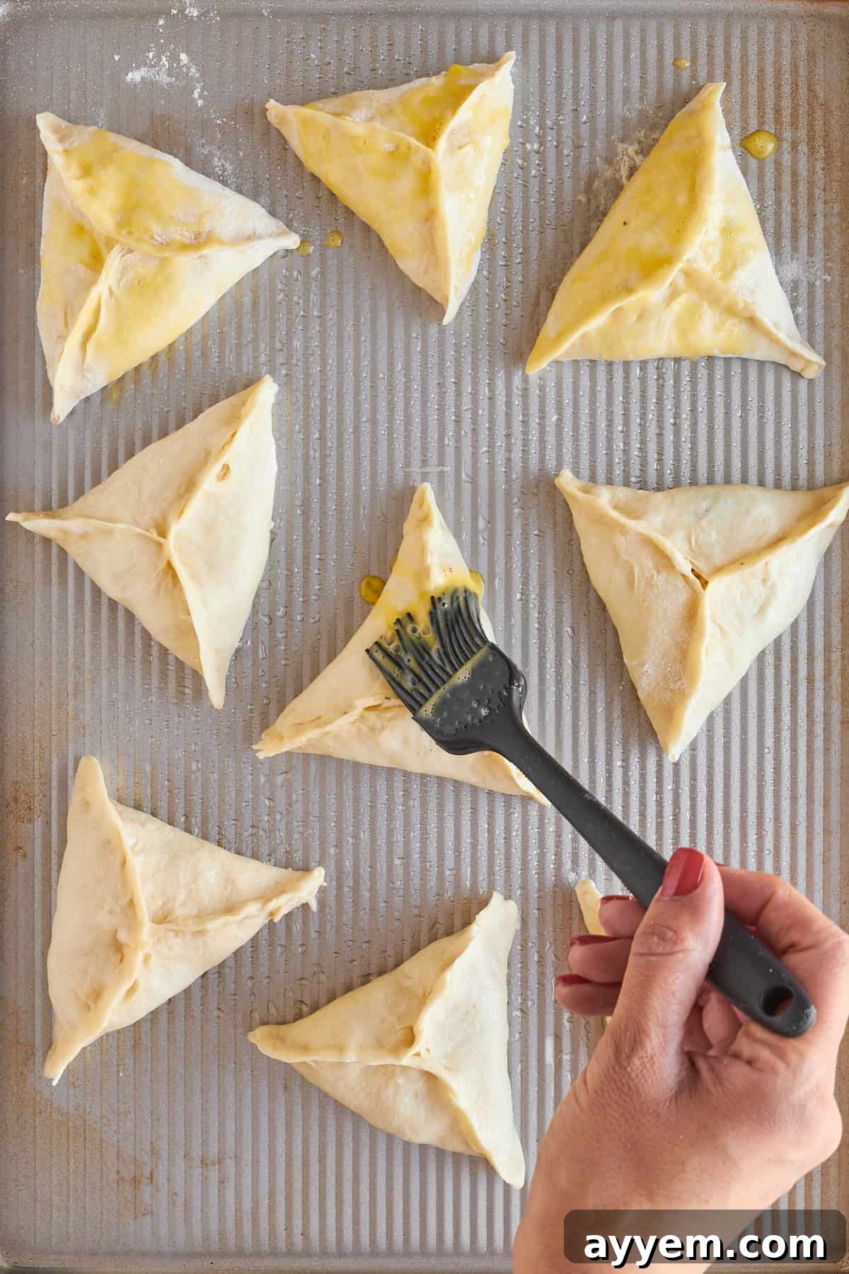 Unbaked fatayer triangles arranged on a parchment-lined baking sheet, glistening with a freshly applied egg wash.