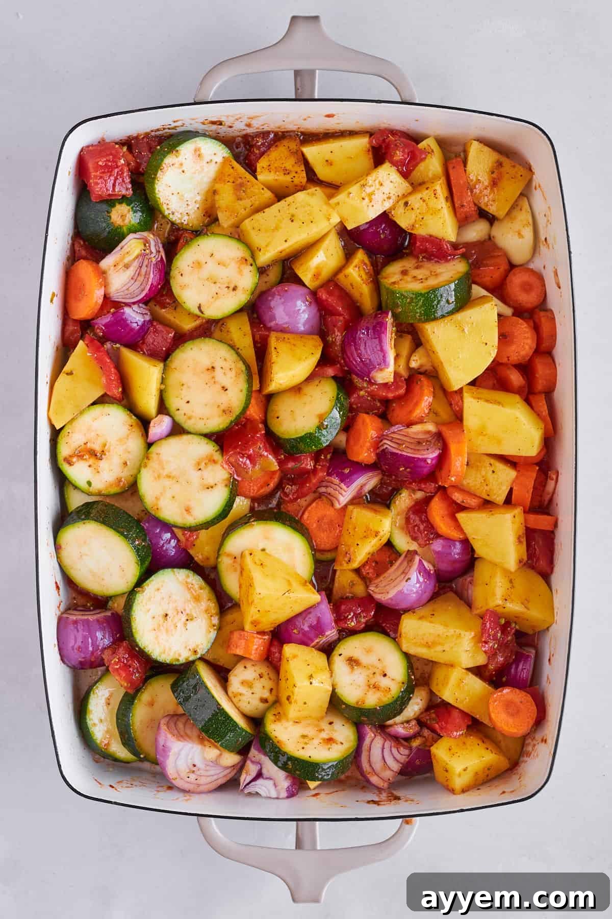 Close-up of seasoned vegetables, including zucchini, potatoes, and red onion, glistening with olive oil and spices in a baking dish.