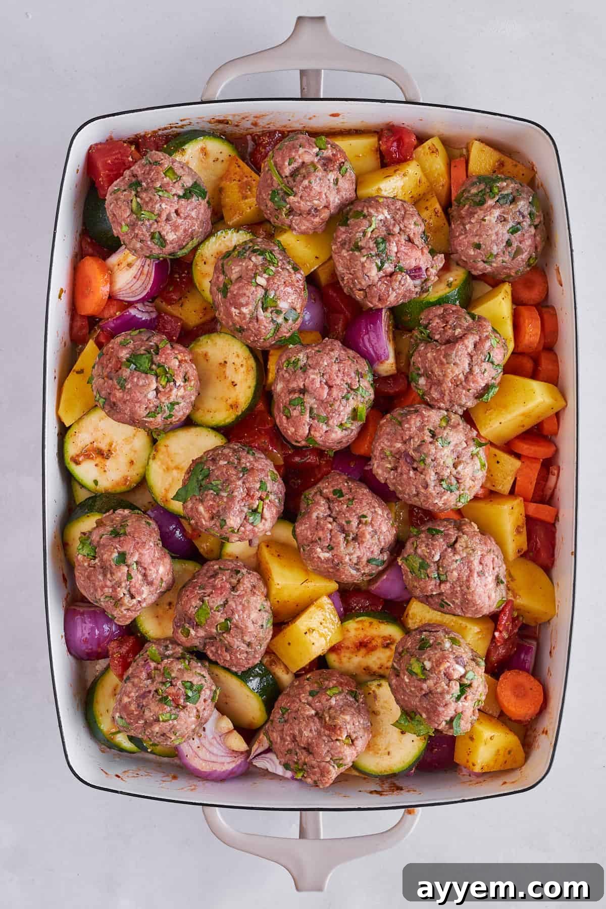 Raw seasoned vegetables topped with perfectly formed raw beef kofta meatballs in a baking dish, ready for baking.